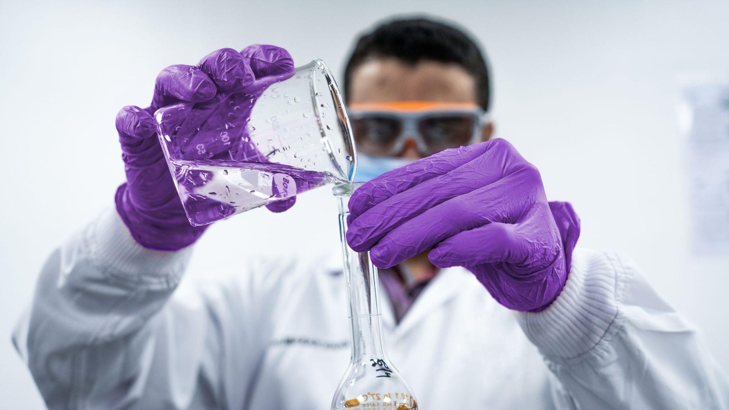 A man wearing purple gloves, white labcoat, and protective glasses, pours clear liquid from a beaker to a flask.