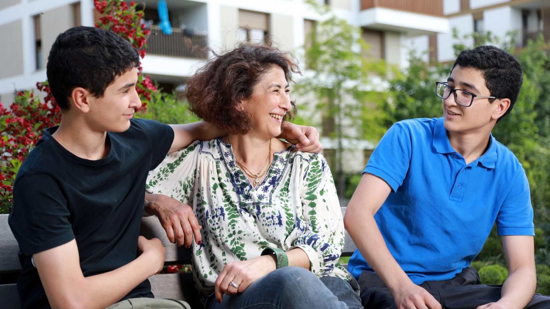A woman sits on a park bench between two teenagers, smiling and talking together in a residential outdoor setting.