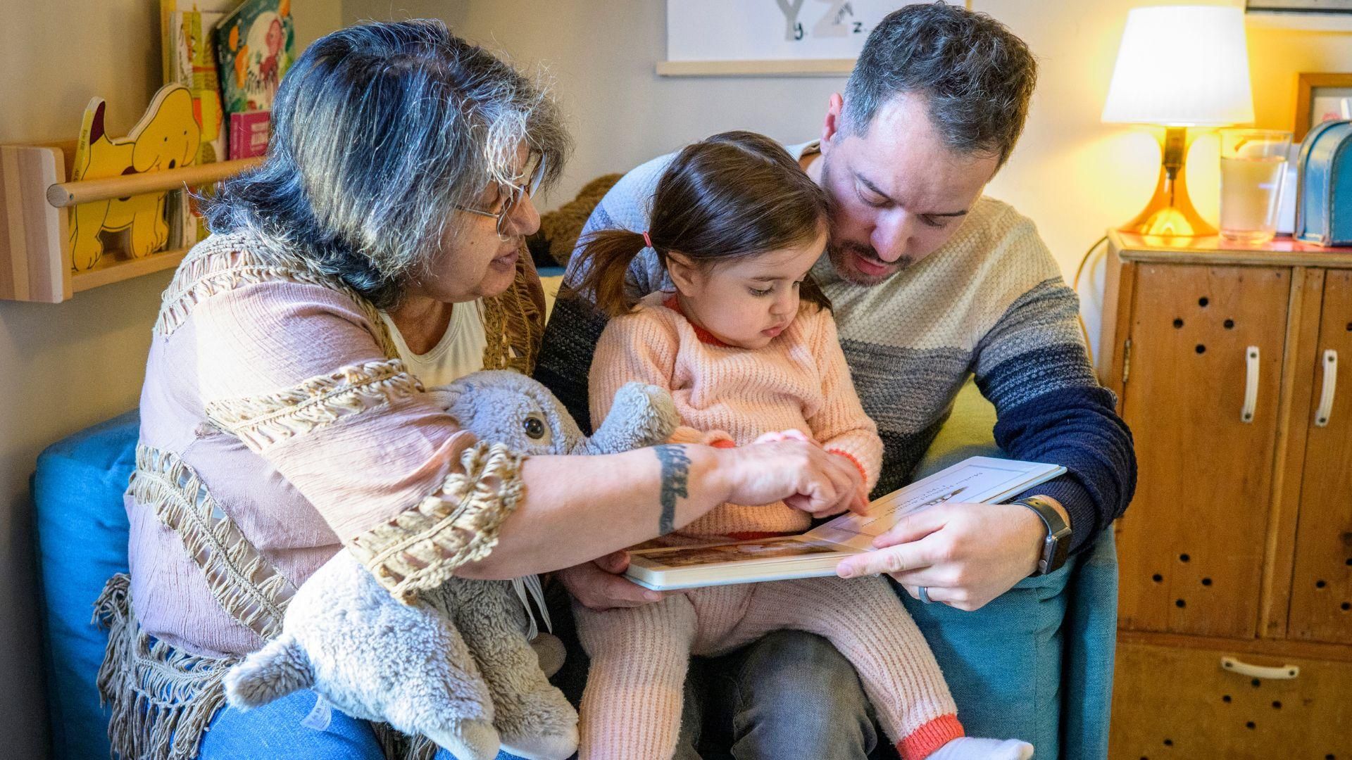 Familia multigeneracional con abuela, padre e hija pequeña leyendo juntos en un ambiente hogareño cálido y protector