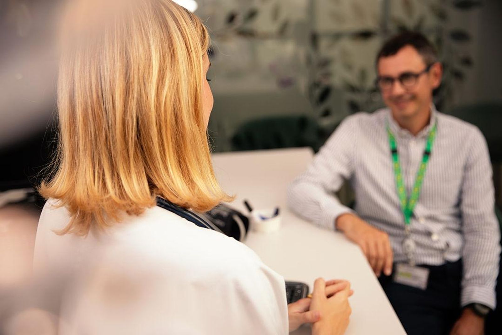 Vista posterior de un profesional de la salud conversando con un paciente con una discapacidad invisible, quien lleva un cordón de girasol para señalar discretamente su necesidad de asistencia o atención, sentado en un escritorio.