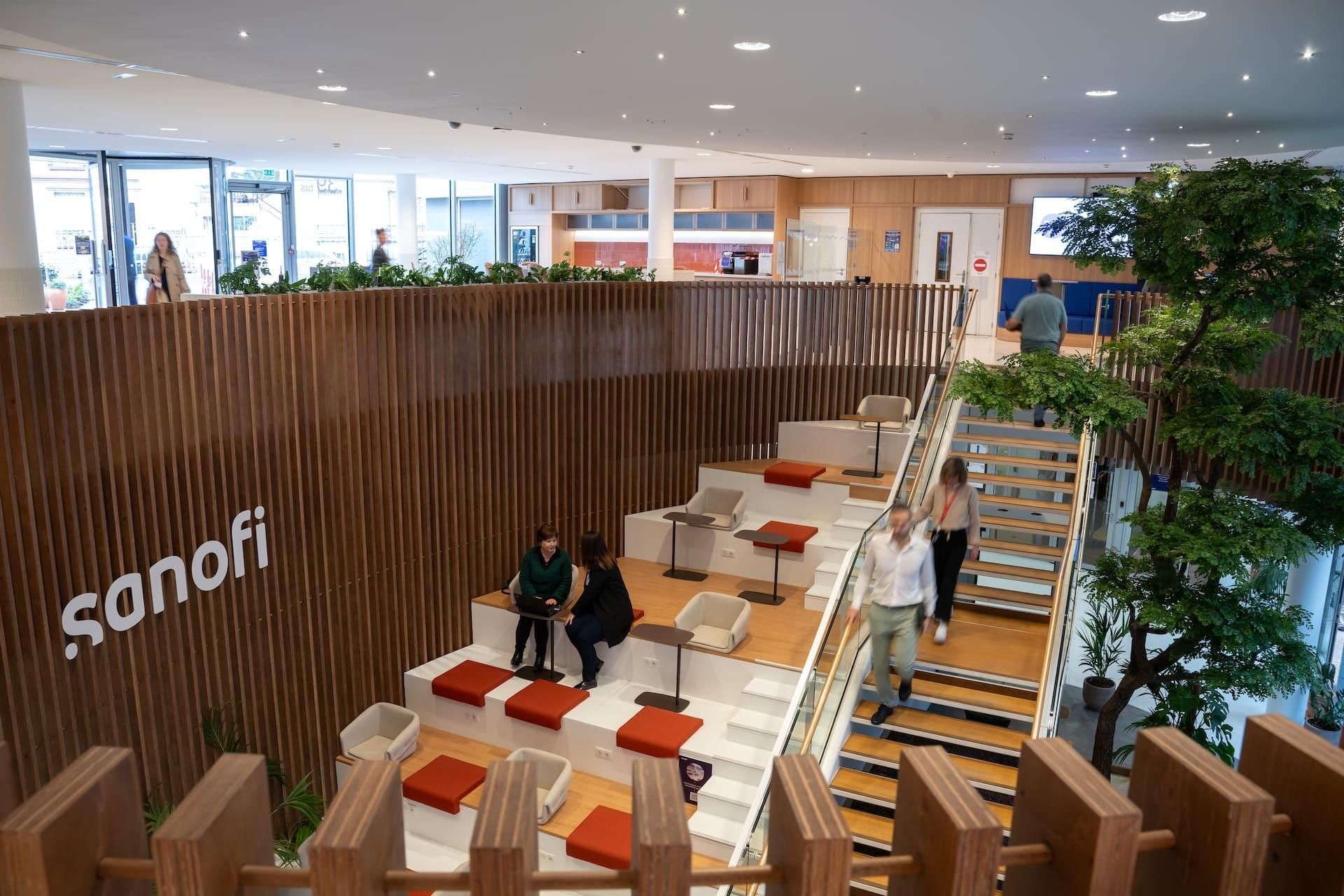 Secondary reception at Sanofi Gentilly office with curved wooden staircase, red seating areas, and employees in a modern collaborative workspace. 