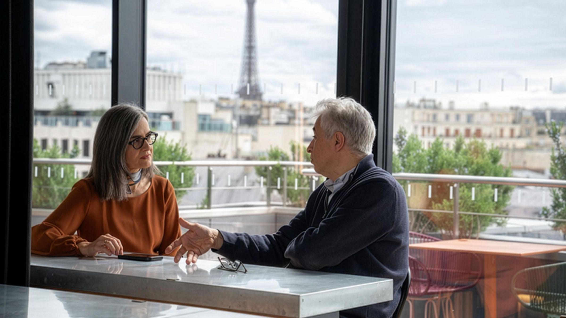 Deux personnes en conversation face à la Tour Eiffel devant de grandes baies vitrées en arrière-plan.