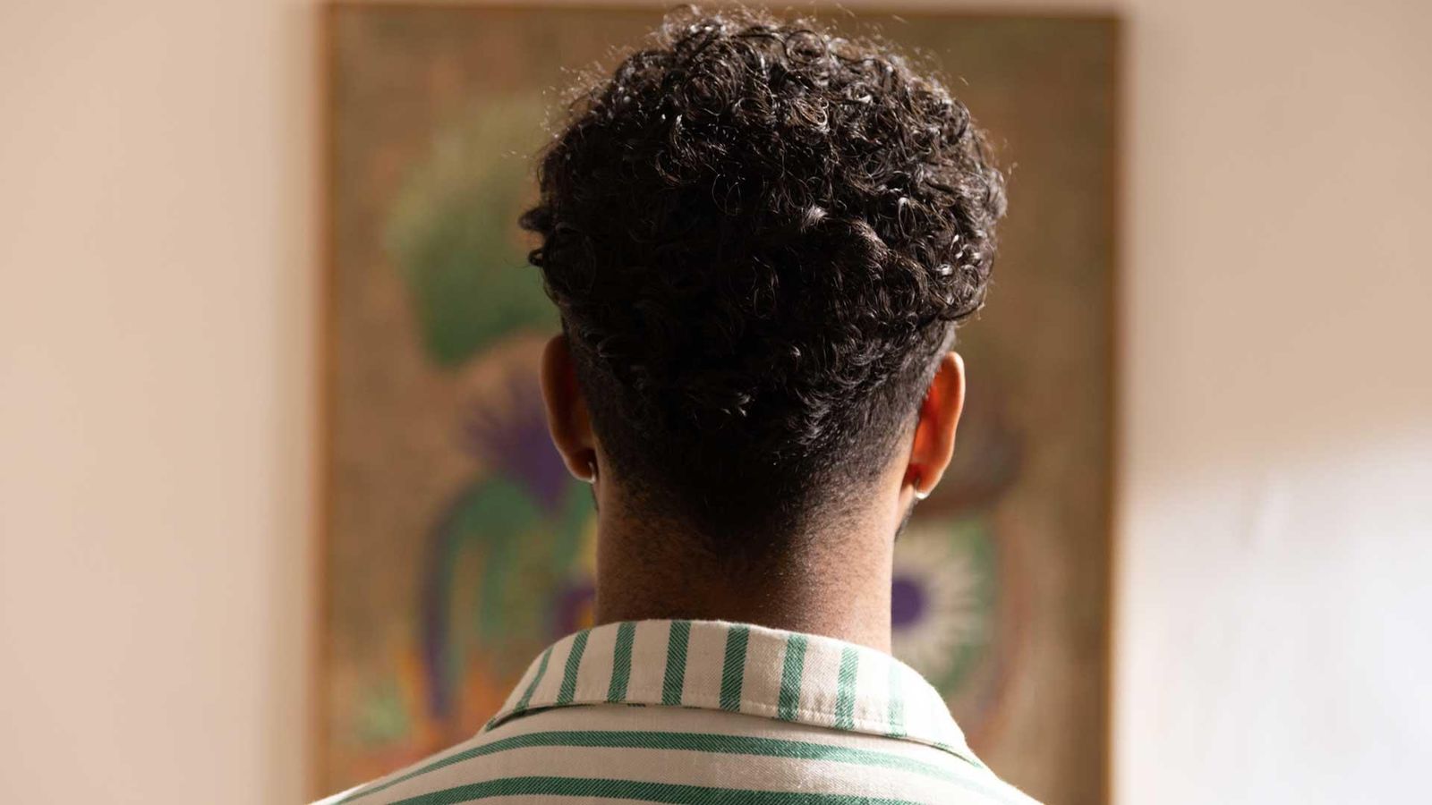 Bruno Araujo de dos, debout avec des cheveux courts bouclés et une chemise rayée blanc et vert, regardant un tableau accroché au mur