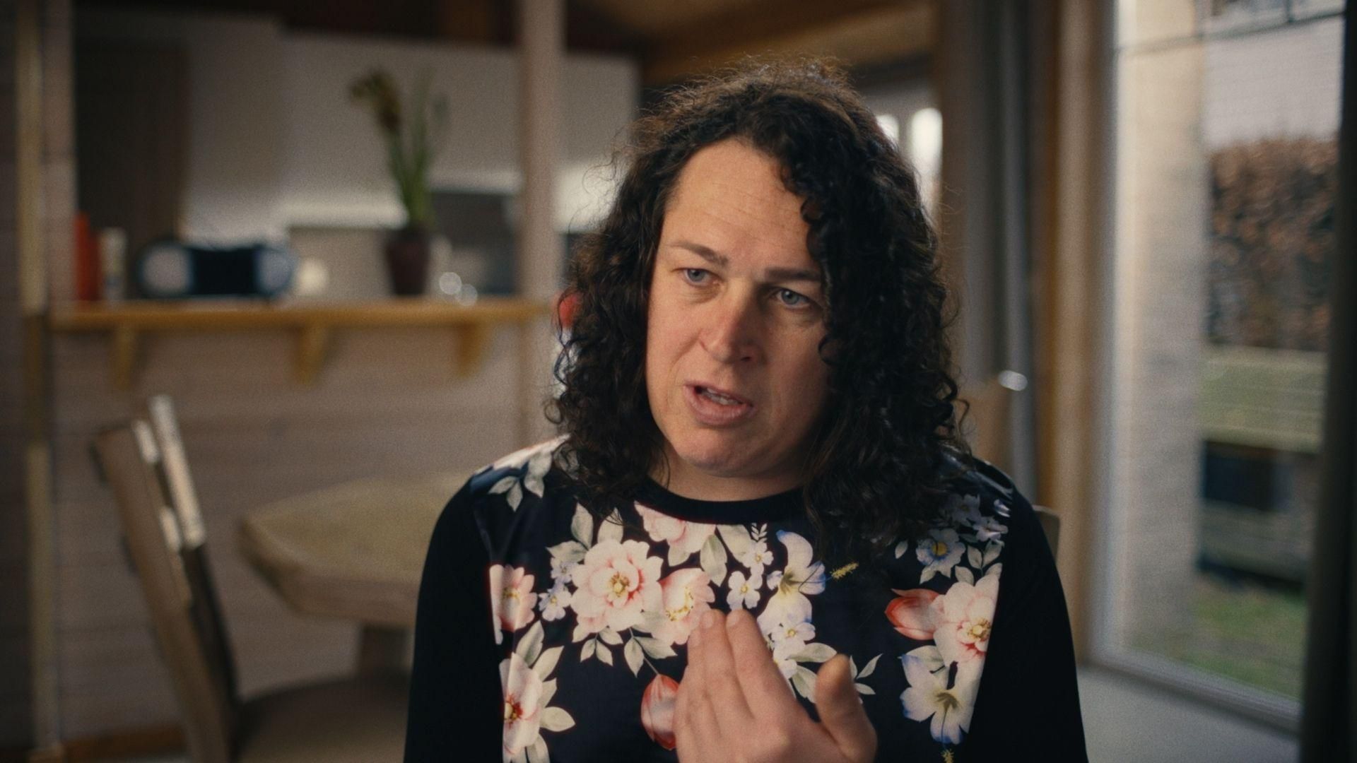 Jill Wuyts de Sanofi Belgique s'exprimant avec animation dans un cadre domestique, portant un haut noir à motifs floraux avec un espace de vie flou en arrière-plan.