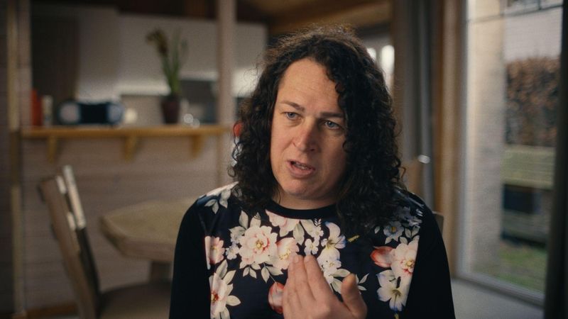 Jill Wuyts de Sanofi Belgique s'exprimant avec animation dans un cadre domestique, portant un haut noir à motifs floraux avec un espace de vie flou en arrière-plan.