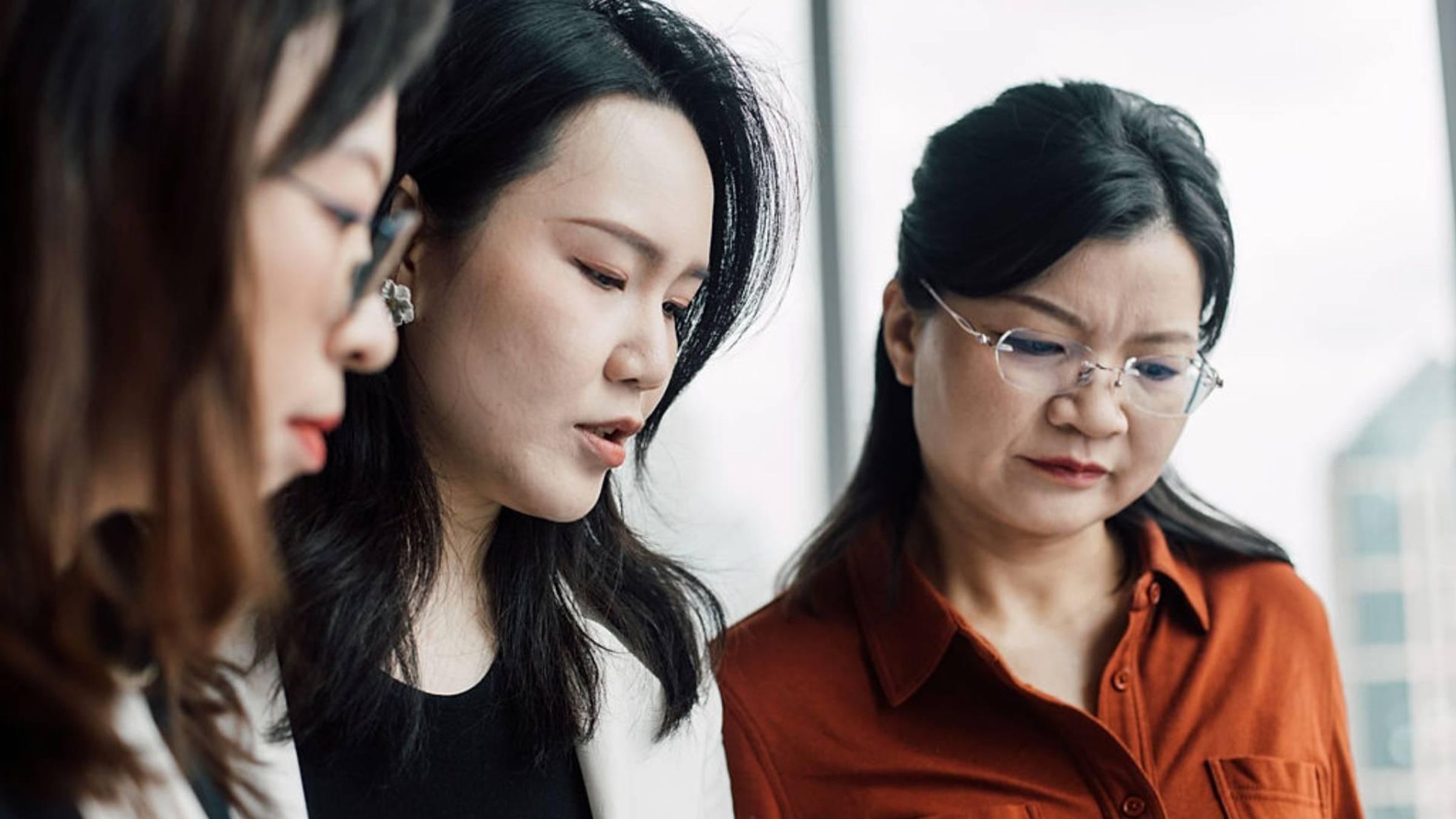 Three Asian women, two wearing glasses, looking at something on the table.