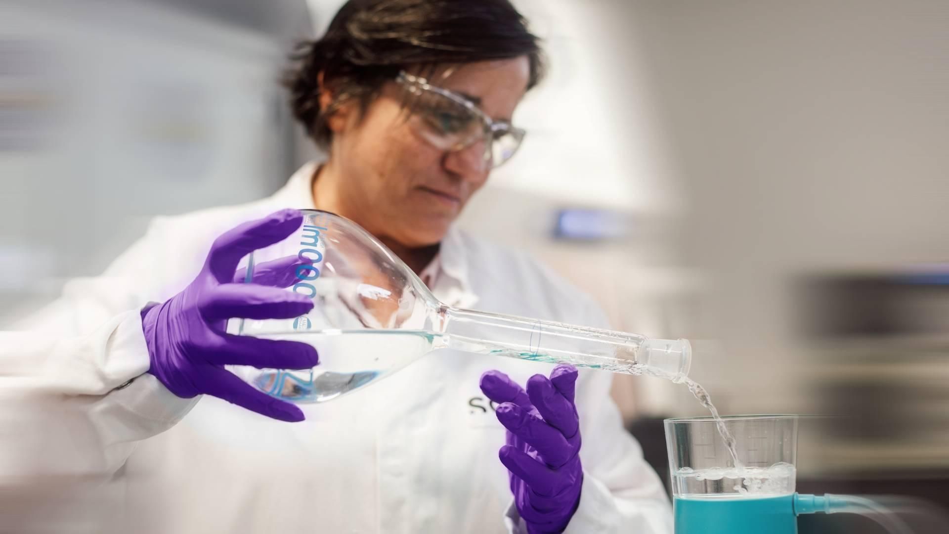 Personne vêtue d'une blouse de laboratoire, de lunettes de sécurité et de gants, versant un liquide dans un dispositif de filtration en laboratoire.