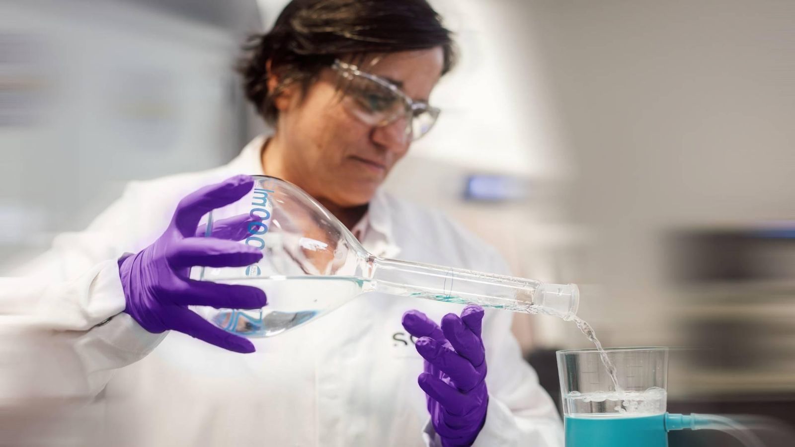 Personne vêtue d'une blouse de laboratoire, de lunettes de sécurité et de gants, versant un liquide dans un dispositif de filtration en laboratoire.