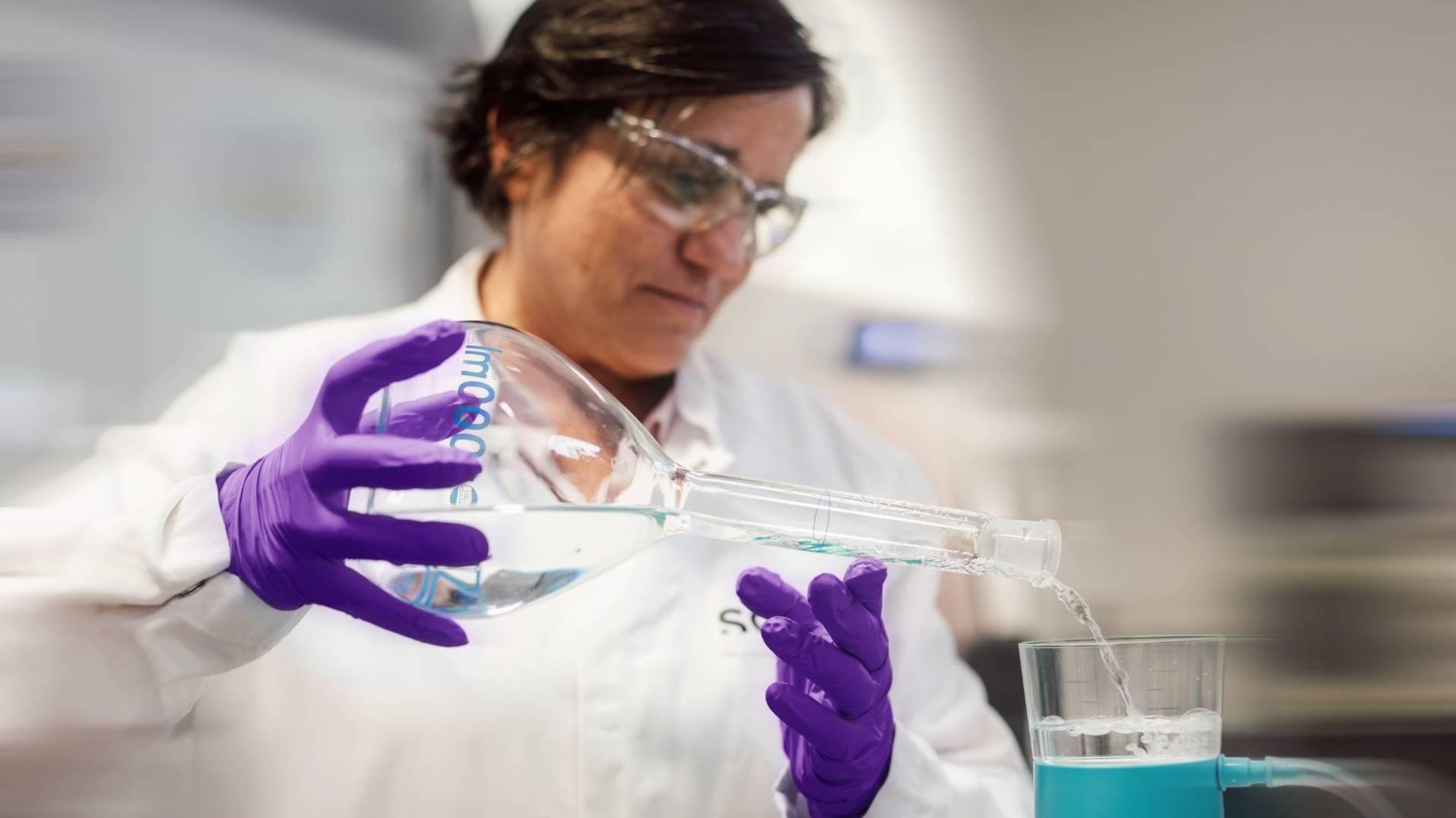 Personne portant une blouse de laboratoire, des lunettes de protection et des gants, versant un liquide dans un dispositif de filtration dans un laboratoire.