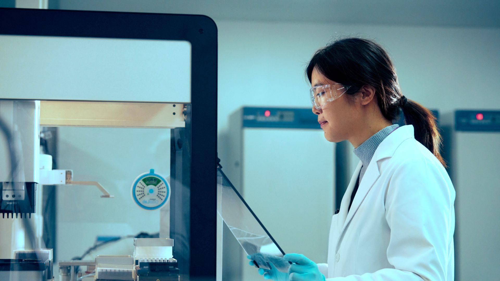 Female scientist in white lab coat and safety equipment working with a tablet at a laboratory workstation with protective hood