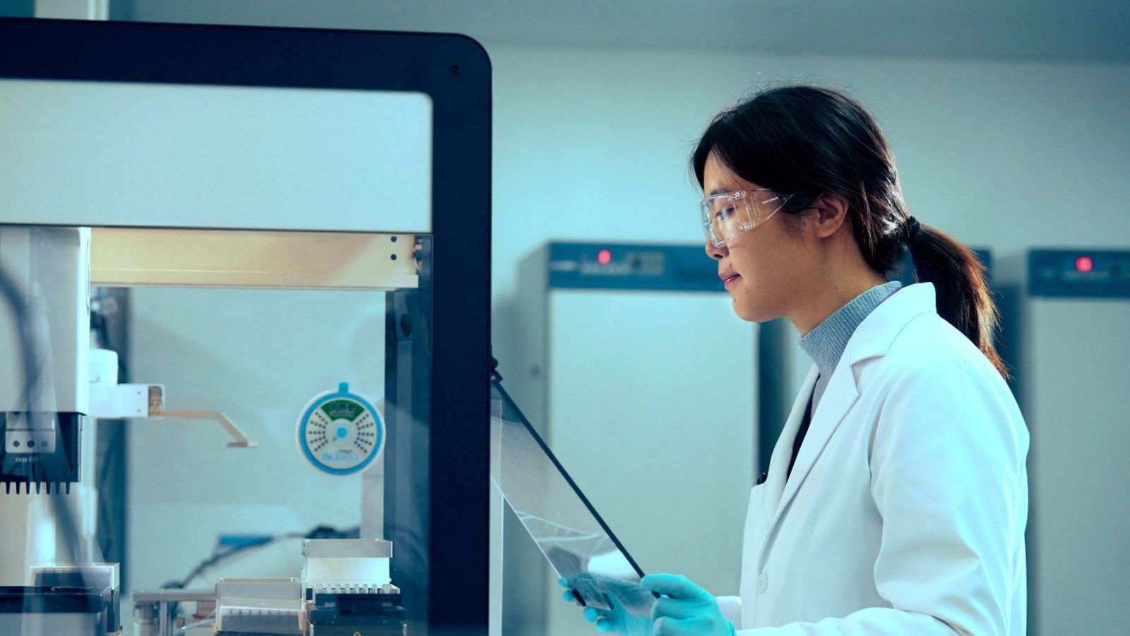 Female scientist in white lab coat and safety equipment working with a tablet at a laboratory workstation with protective hood