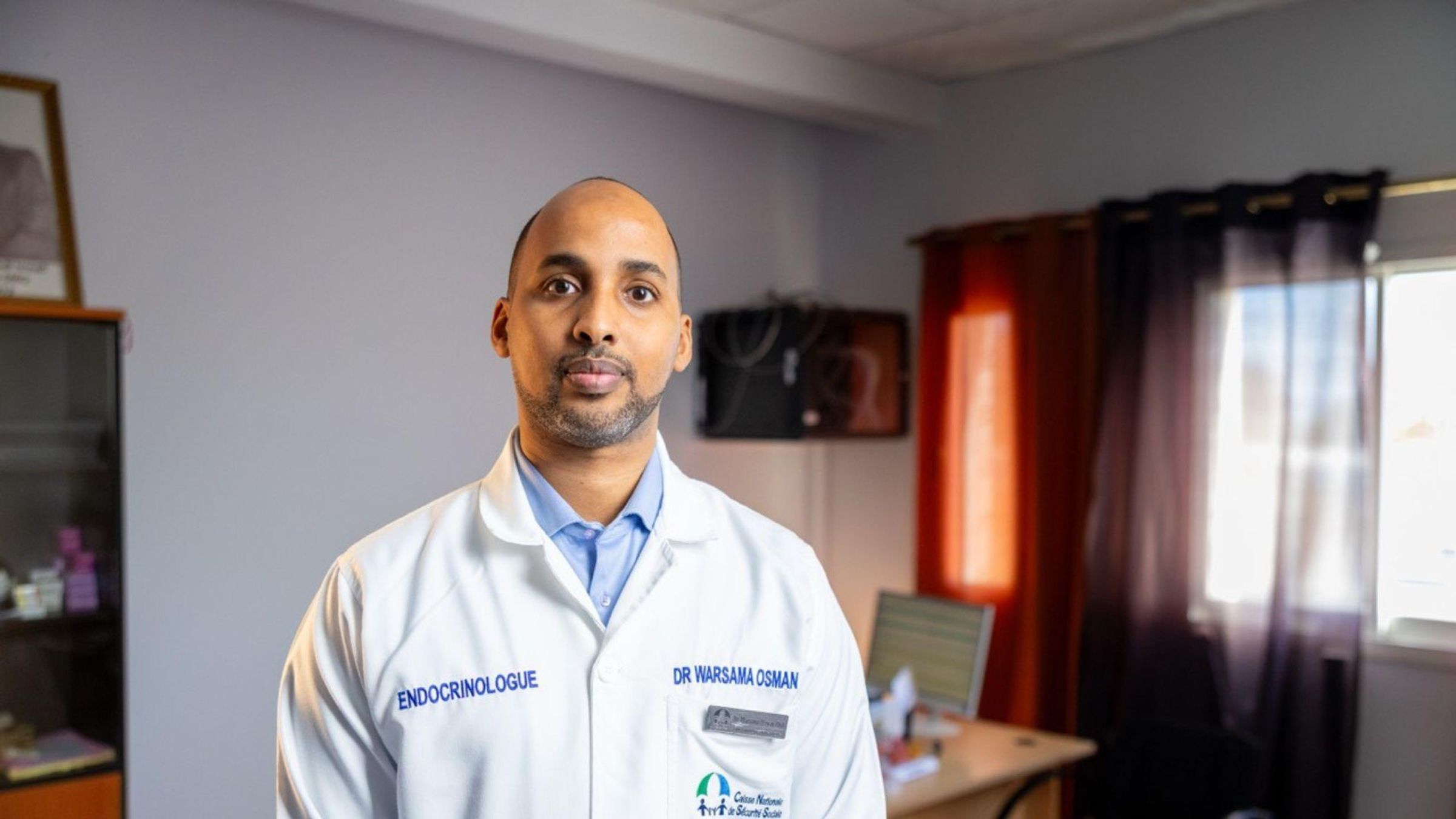 Dr Warsama Osman, endocrinologue, debout dans son cabinet médical portant une blouse blanche avec son nom et sa spécialité affichés