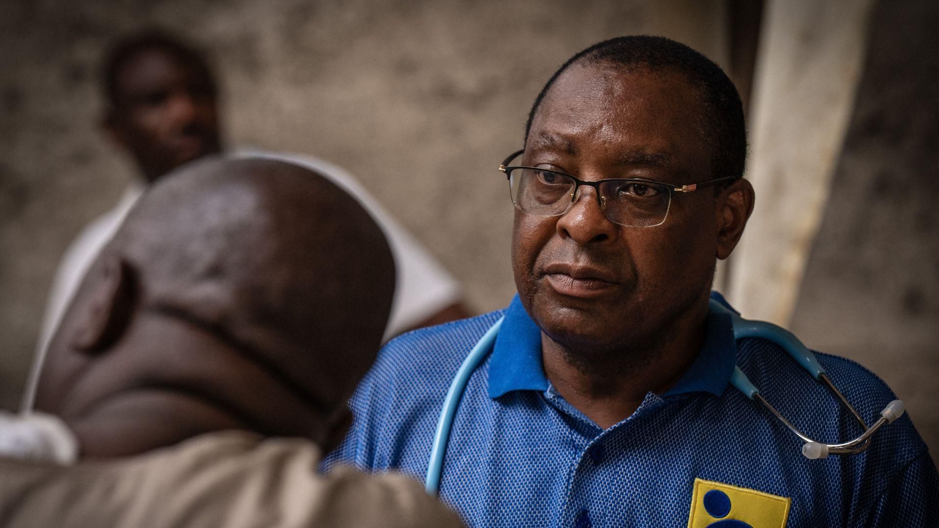 Le Dr. Yohana Mokiwa, avec une expression sérieuse, chemise bleue et stéthoscope, s'entretient avec un patient dans une clinique éphémère