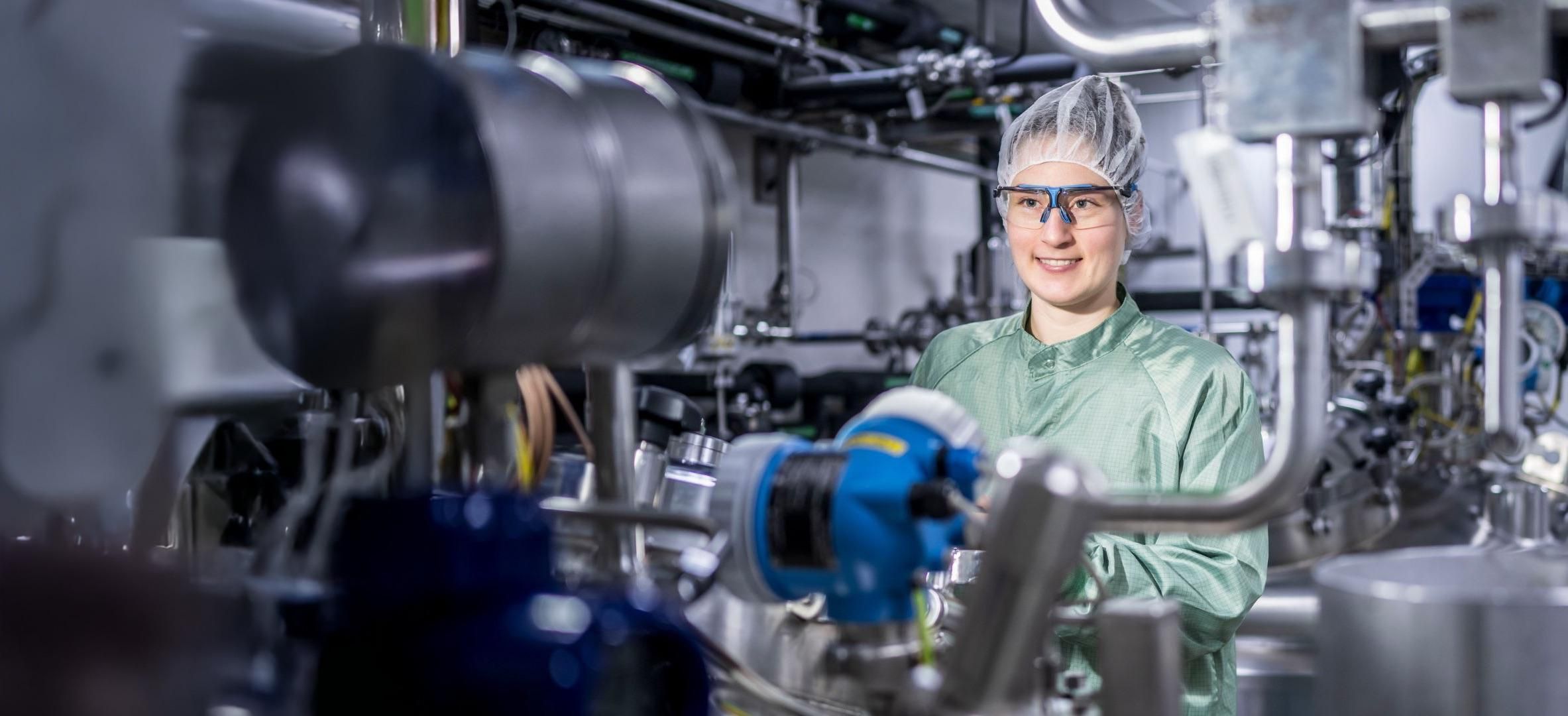 Julia Schwendner, Process Responsible Scientist, smiling in a green lab coat, hairnet and safety goggles while standing in front of industrial bioprocessing equipment at Sanofi's Frankfurt facility.