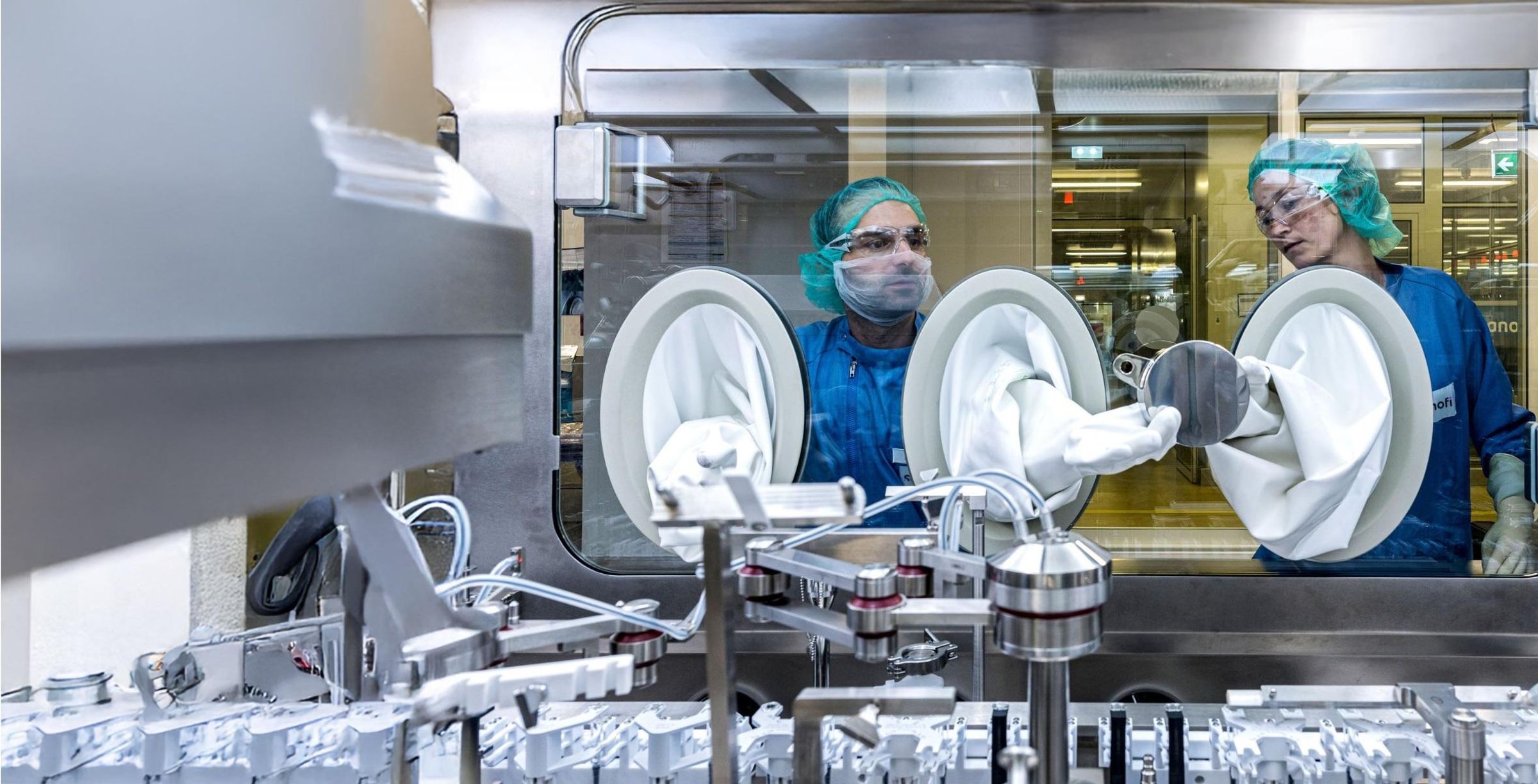 Emre Canaki and Stefanie Kutschmann pass a metal disk between them by way of gloveboxes inside a large steel machine.