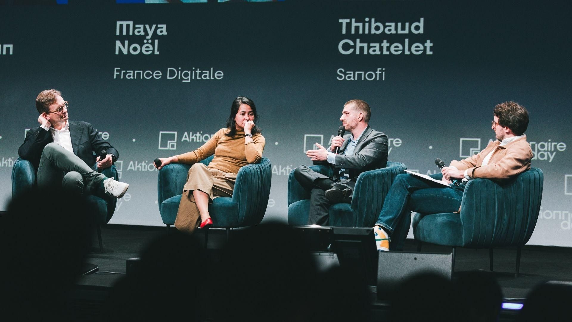 Four panelists engaged in discussion on stage at Aktionnaire Day conference, seated in teal chairs. From left to right: a man in dark suit with glasses, a woman in beige top and brown skirt with red heels, a man in dark suit, and a man in light brown jacket with glasses.