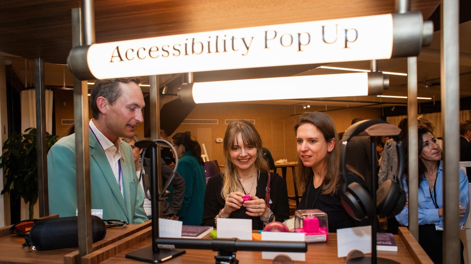 Un groupe de personnes rassemblées autour d’un stand intitulé « Accessibility Pop Up », présentant divers dispositifs d’accessibilité comme des écouteurs et des objets tactiles, dans un espace événementiel bien éclairé.