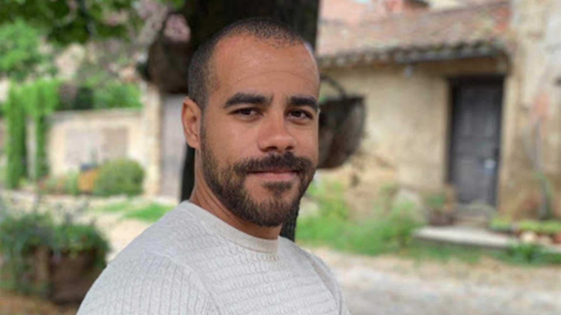 Homme barbu en chemise claire debout à l'extérieur devant un ancien bâtiment en pierre avec porte en bois, entouré de verdure luxuriante