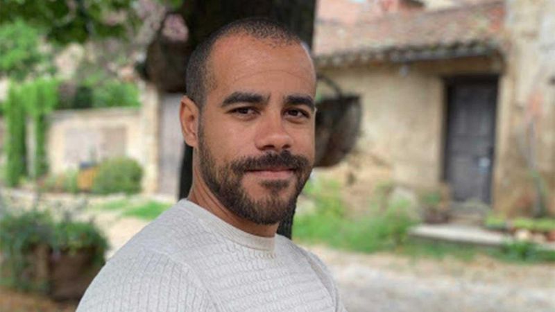 Homme barbu en chemise claire debout à l'extérieur devant un ancien bâtiment en pierre avec porte en bois, entouré de verdure luxuriante