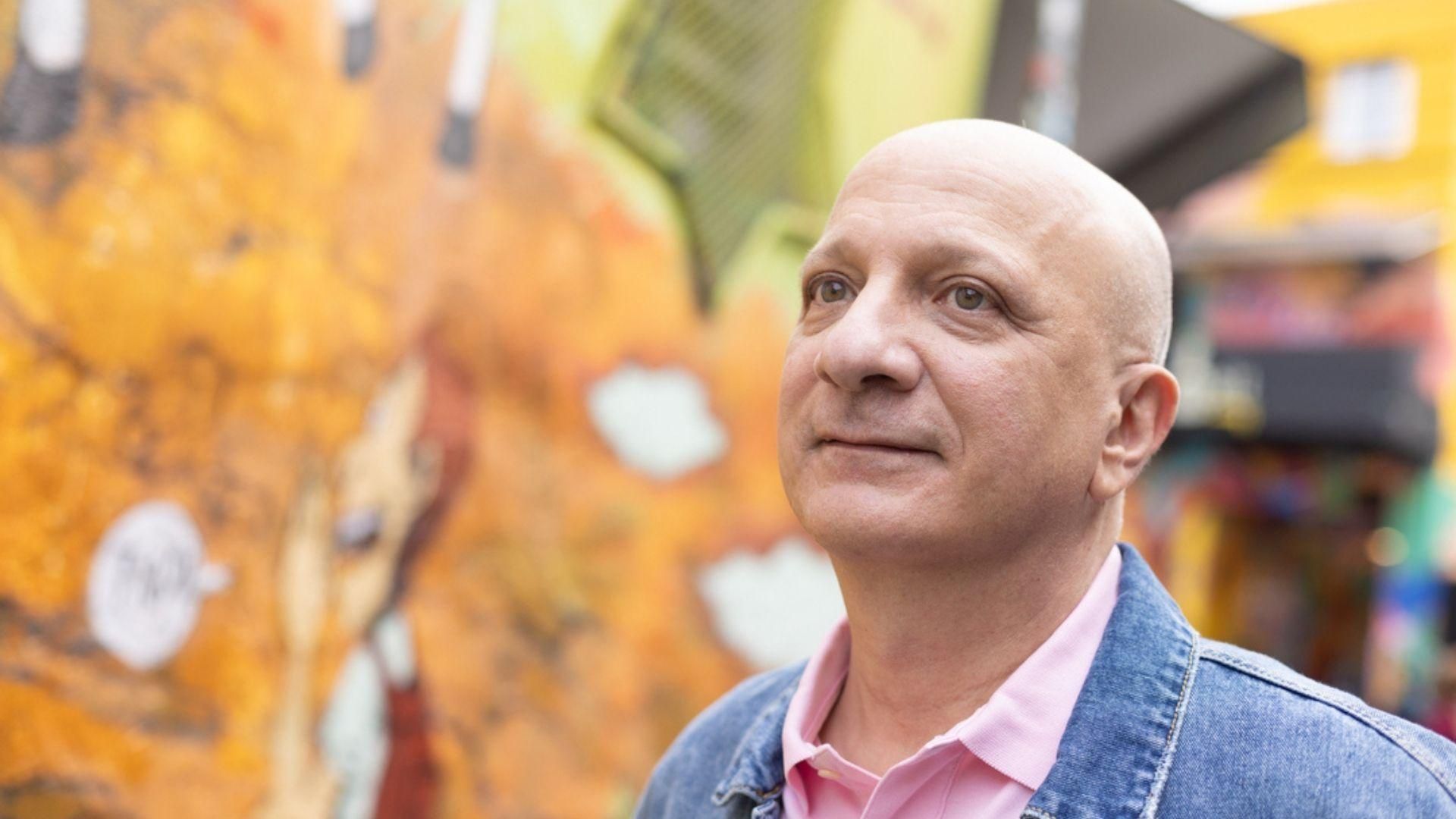 Pedro Pittella, au crâne rasé, portant une chemise rose et une veste en jean, se tient devant un mur coloré à São Paulo