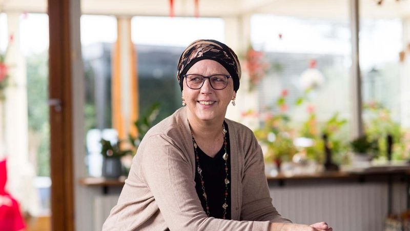 Une femme assise à l'intérieur, portant un cardigan beige et un foulard sur la tête, souriant à la caméra. Derrière elle se trouvent une porte vitrée, des étagères avec des plantes et une fenêtre