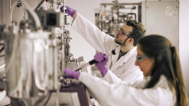 Deux scientifiques, un homme et une femme, travaillant dans un laboratoire. Tous deux portent des blouses blanches. L'homme a un bandana et des lunettes, la femme porte des gants. Des équipements de laboratoire sont visibles en arrière-plan.