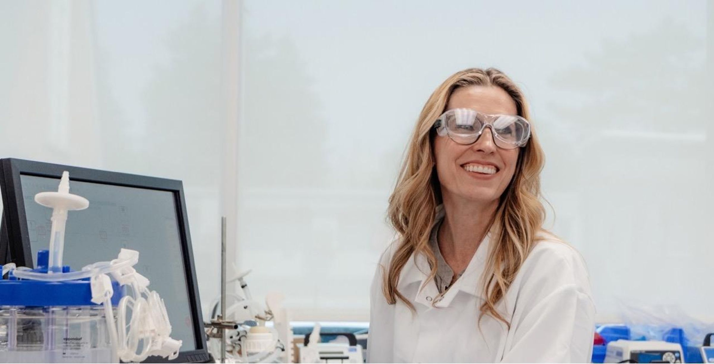 Female, Senior Unit Head, Drug Product Development, wearing a white lab coat and safety glasses and purple gloves, smiles while working at her lab computer