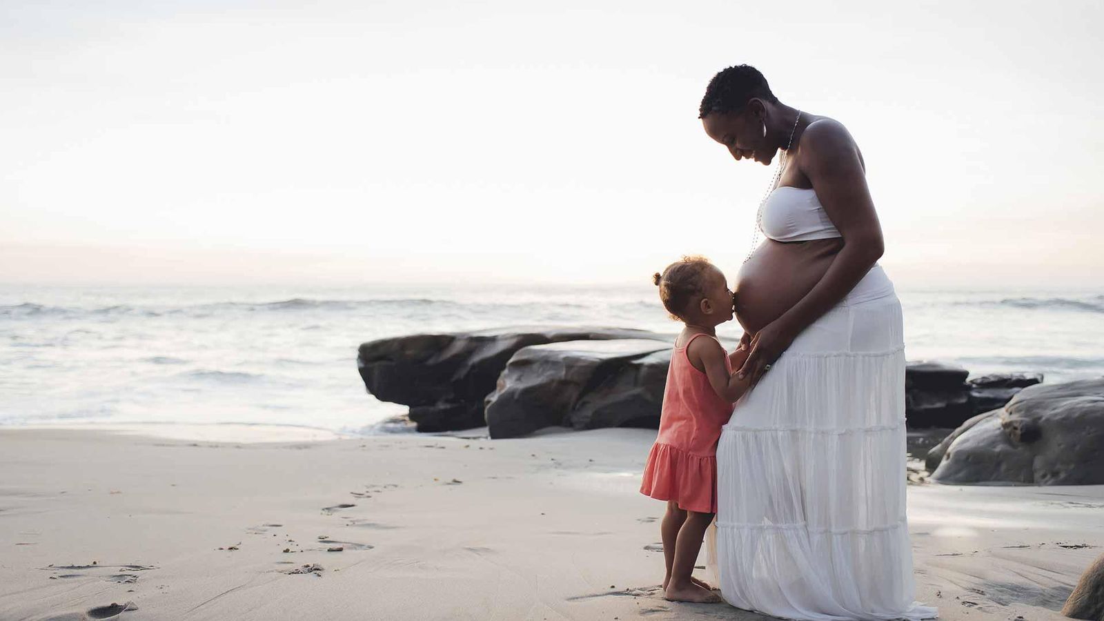 Fille embrassant le ventre de sa mère enceinte sur la plage au coucher du soleil 