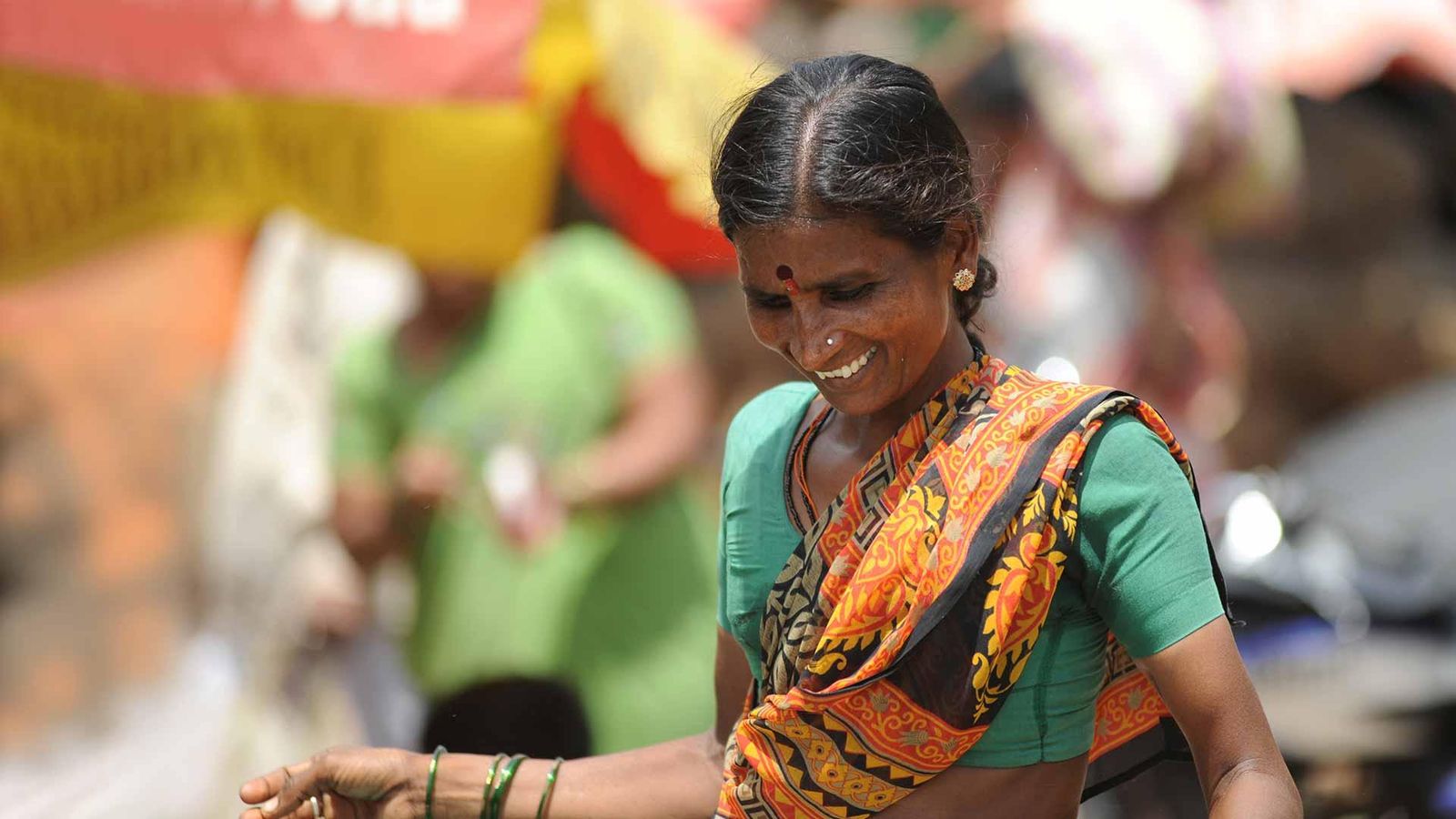 Portrait d'une femme indienne souriante portant un chemisier vert et un dupatta orange et noir, ornée de bracelets verts et d'un bindi, avec d'autres personnes en arrière-plan flou lors d'un événement en extérieur