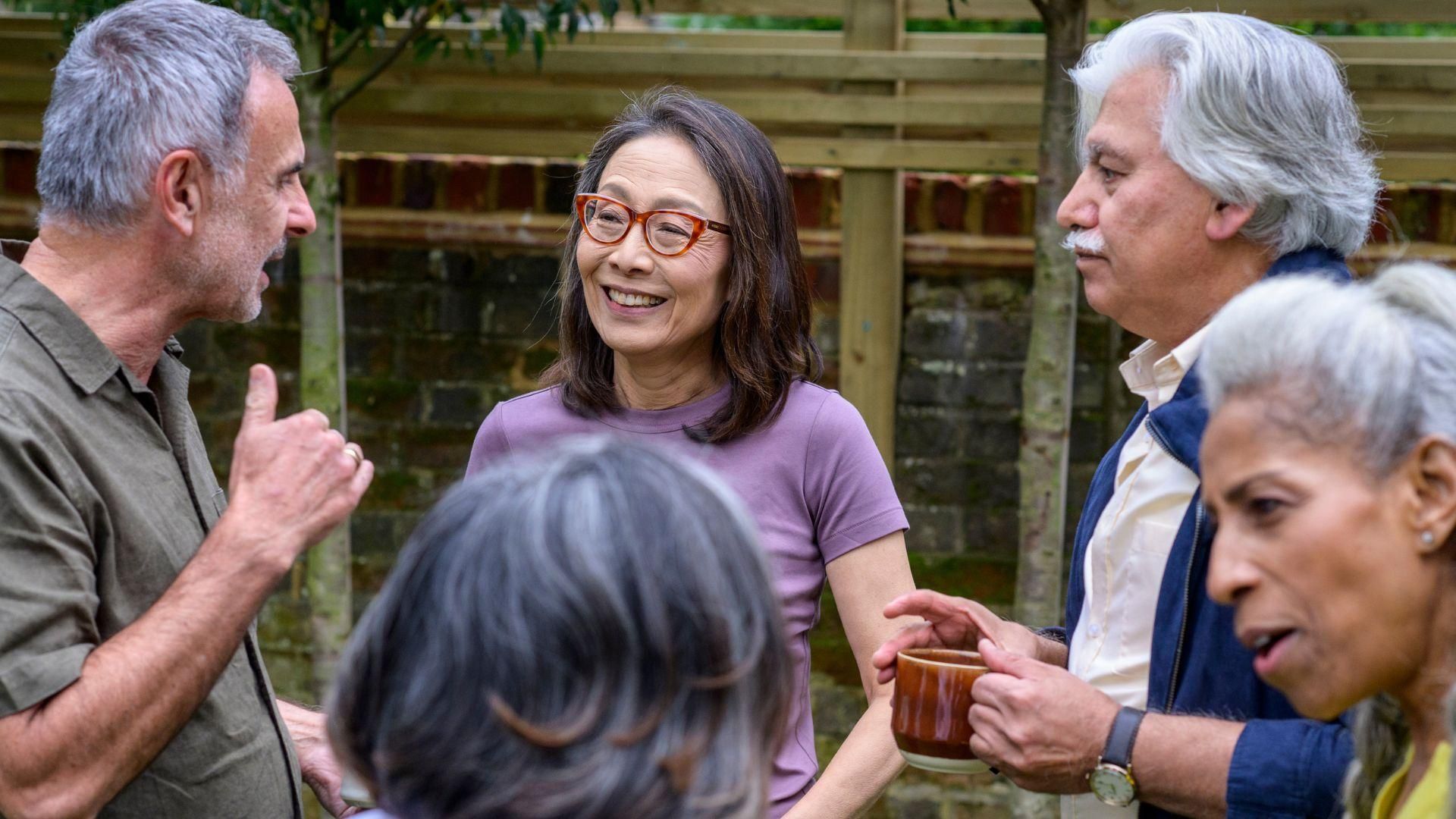 A group of older adults standing in a garden, talking, with a wooden fence and greenery in the background.