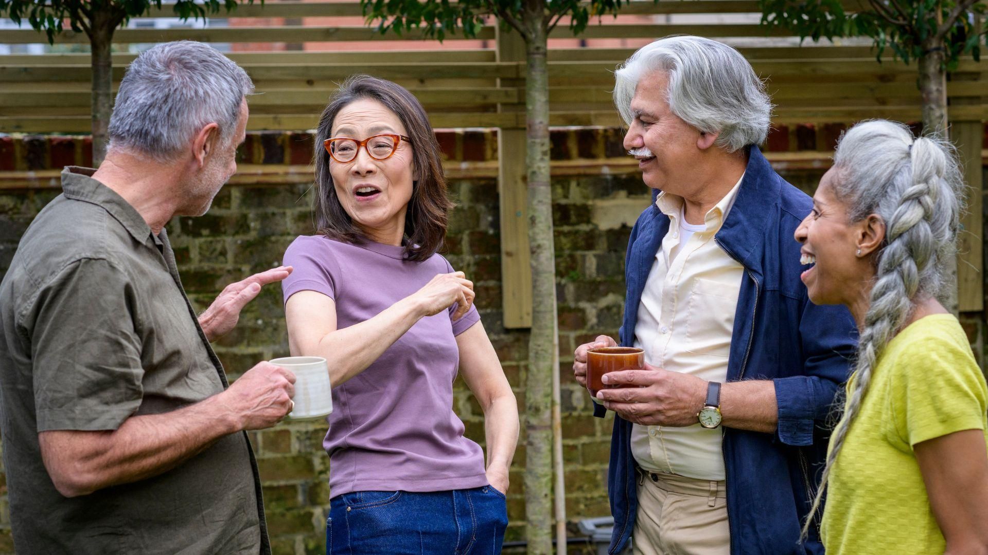 A group of older adults standing in a garden, talking and holding mugs, with a wooden fence and trees in the background.