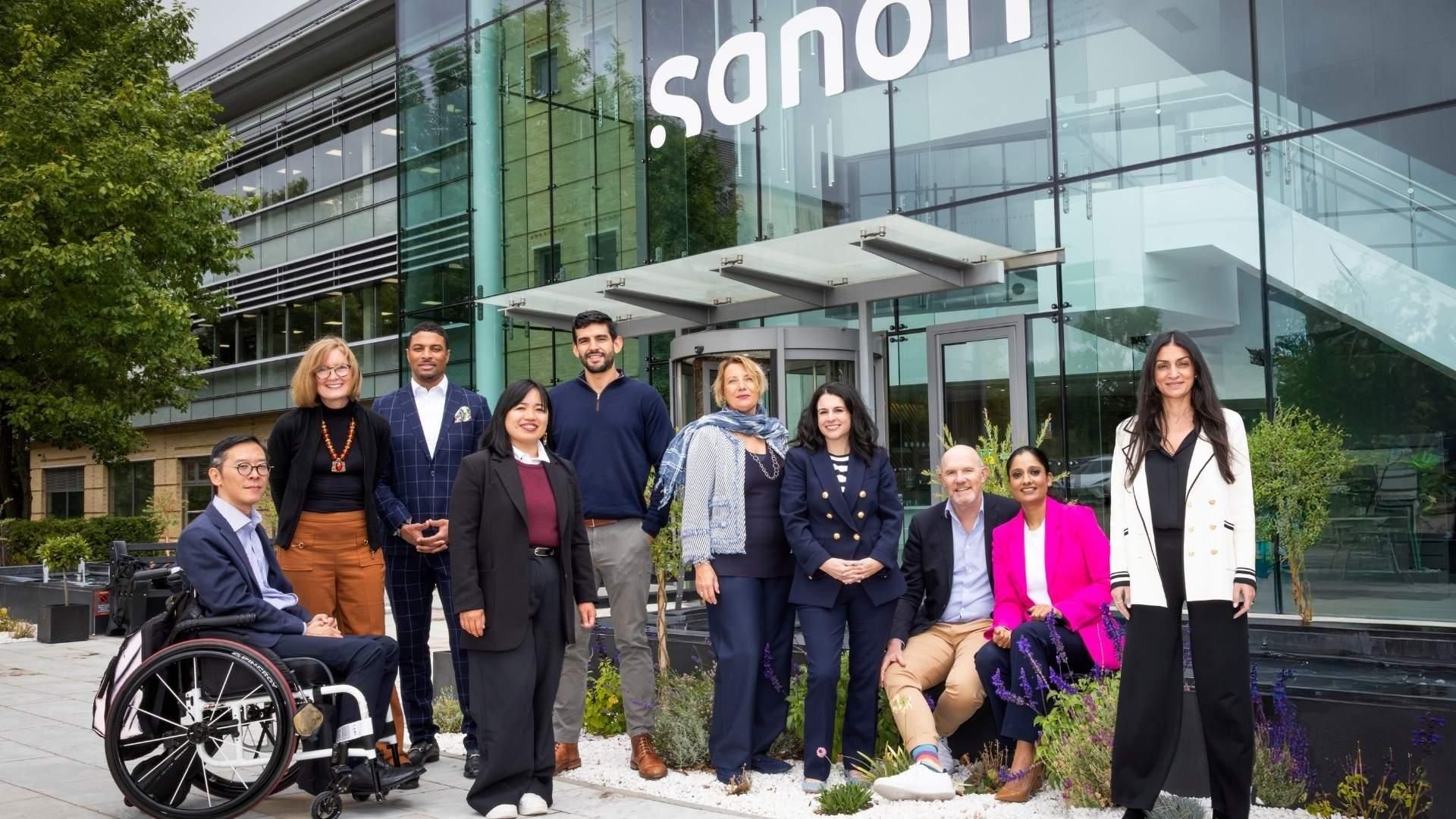 Un groupe diversifié de dix professionnels en tenue décontractée (business-casual) pose pour un portrait devant un bâtiment moderne en verre arborant le logo Sanofi.
