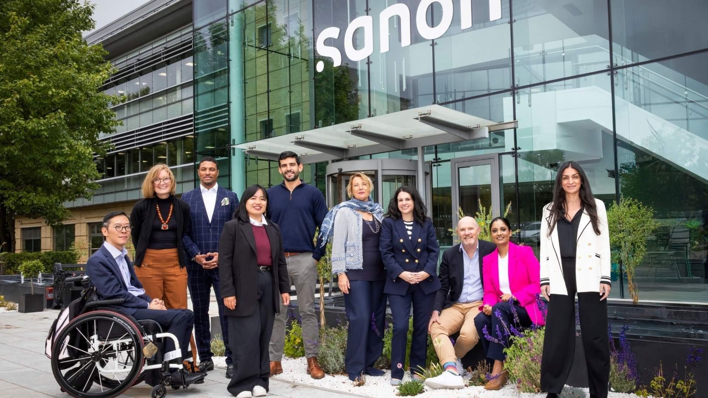 Un groupe diversifié de dix professionnels en tenue décontractée (business-casual) pose pour un portrait devant un bâtiment moderne en verre arborant le logo Sanofi.