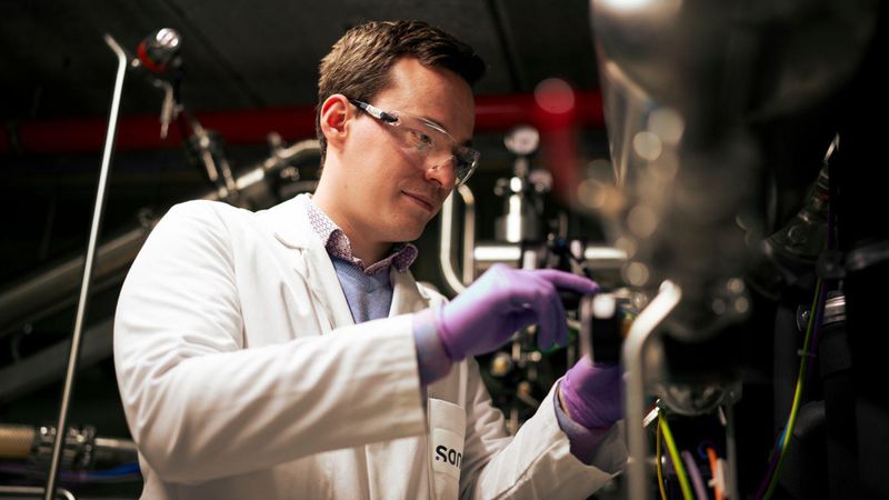 Anthony Van Poucke is wearing a white lab coat and purple gloves, and is adjusting a dial on a wall of steel tubes and valves