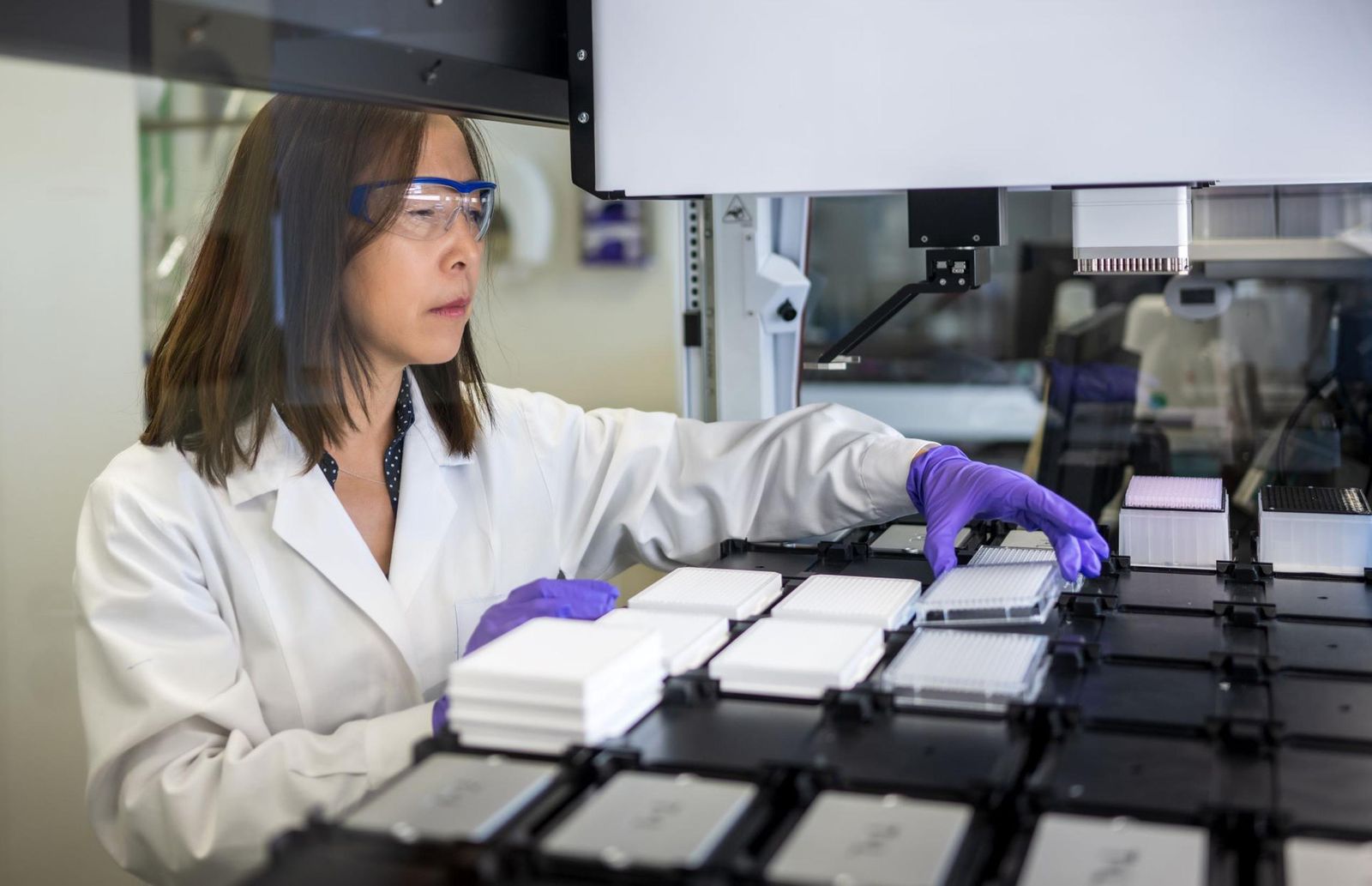 Ziyu Li inspecte les échantillons sur une machine de laboratoire, en portant une blouse de laboratoire blanche, des gants violets et des lunettes de sécurité.